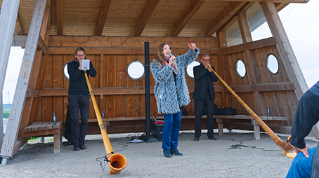 Zwei Männer die Alphorn spielen, eine Frau in der Mitte singt dazu